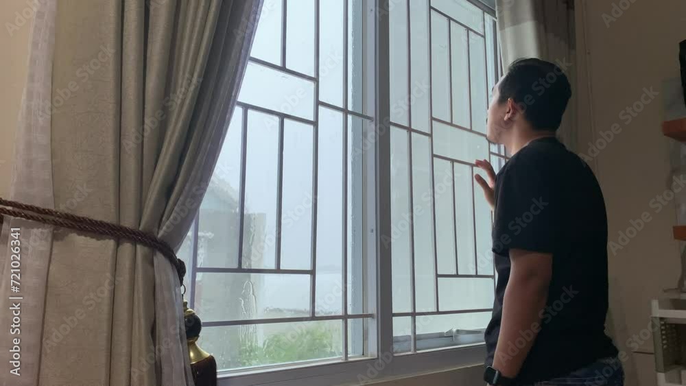 Pensive sad asian man stands at the window. It is raining outside.