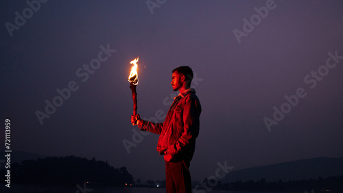 Image of brave man holding burning stick while moving in darkness. Young man holding a fire stick on the mountain top near the sea. 