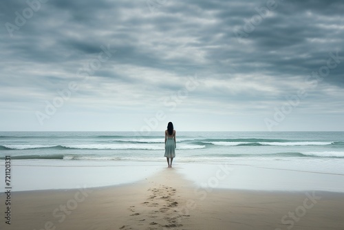 A lone person stands facing the ocean, reflecting in solitude on a vast, calm beach under an overcast sky.