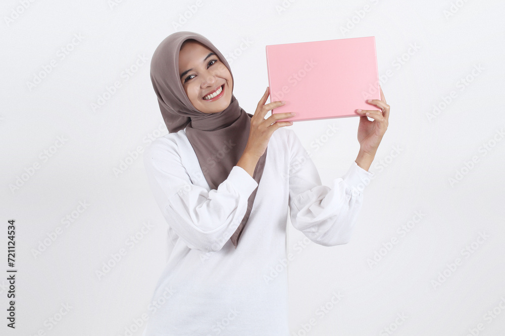 A beautiful Asian Muslim woman is holding a pink gift over her shoulder while smiling showing her teeth A beautiful Asian Muslim woman is holding a pink gift over her shoulder while smiling showing her teeth