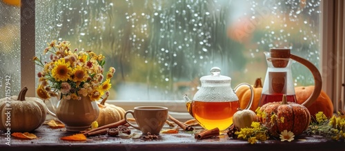 Cozy autumn display with hot tea, honey, pumpkins, cinnamon, and flowers by rainy window.