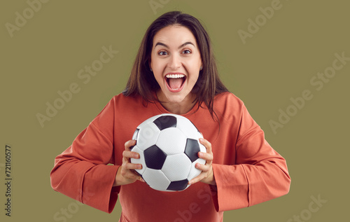 Portrait of funny soccer fan woman who is happy with crazy and happy expression on her face. Young Caucasian woman with football ball in hands joyfully shouting isolated on khaki background. © Studio Romantic