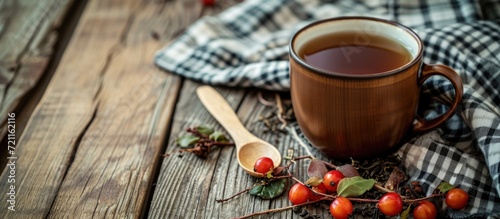 Enamel mug with herbal tea, dried rosehips, wooden spoon, checkered napkin on wooden table.