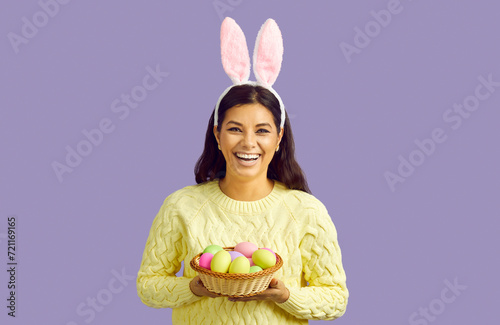 Cheerful woman in rabbit ears holding wicker bowl with Easter eggs on pastel purple background. Portrait of joyful woman with soft Easter bunny ears and in yellow sweater laughing looking at camera.