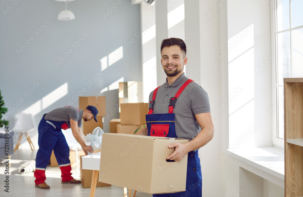Portrait of happy worker from professional man and van moving delivery service. Young male loader in uniform workwear standing in living room, holding cardboard box, looking at camera and smiling Portrait of happy worker from professional man and van moving delivery service. Young male loader in uniform workwear standing in living room, holding cardboard box, looking at camera and smiling
