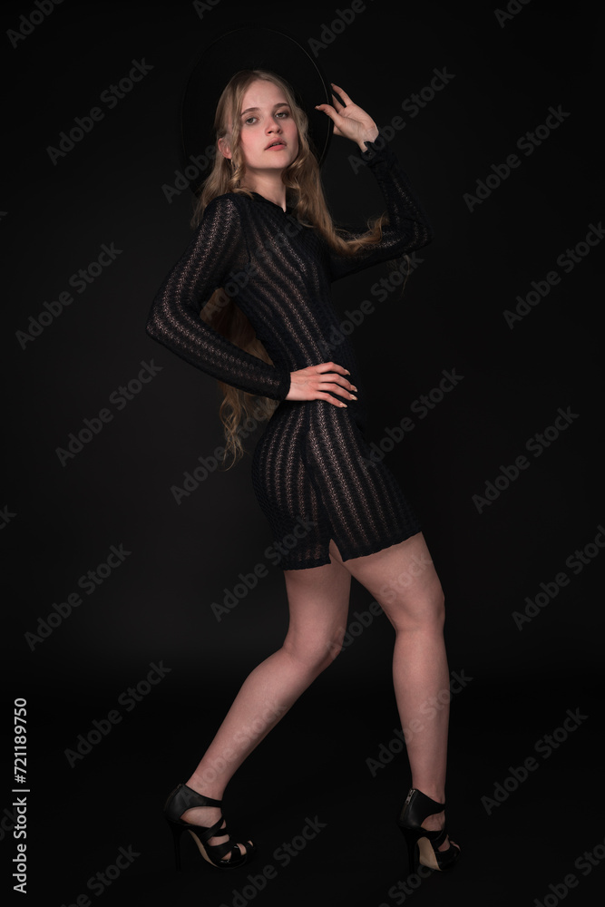Blonde fashion model looks amazing in black short knitted translucent dress, wide-brimmed felt hat, high heels. Full length portrait captures her from three-quarter rear view against black background Blonde fashion model looks amazing in black short knitted translucent dress, wide-brimmed felt hat, high heels. Full length portrait captures her from three-quarter rear view against black background