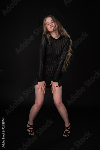 Confident young woman exuding elegance and style. With her eyes closed and lower lip pursed. She is wearing black short knitted translucent dress, wide-brimmed felt hat and high-heeled shoes