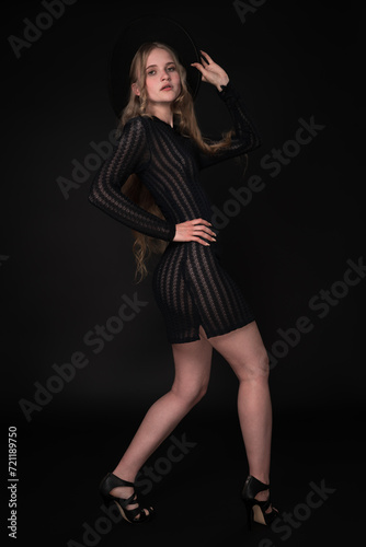 Blonde fashion model looks amazing in black short knitted translucent dress, wide-brimmed felt hat, high heels. Full length portrait captures her from three-quarter rear view against black background