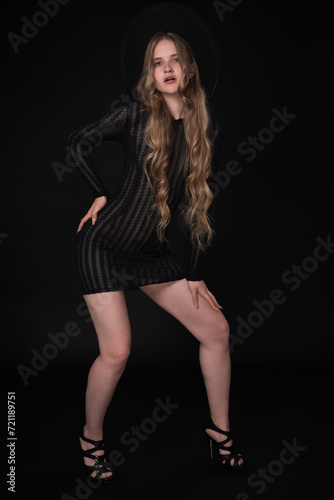 Full length of young woman with long legs, posing on black background. Blonde model with long hair is wearing black short knitted transparent tight dress, wide brimmed felt hat and high heeled shoes
