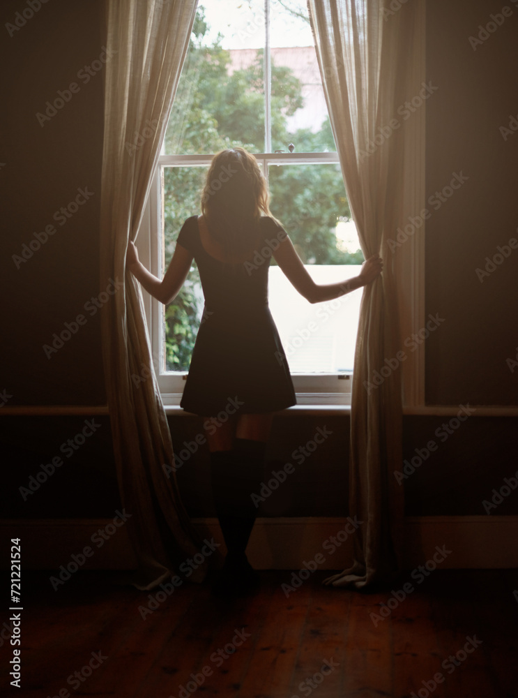 Woman, curtain and morning by window for sunlight, vitamin D or sunshine in bedroom at home. Rear view of female person standing by blinds or glass in casual clothing and looking out in neighborhood Woman, curtain and morning by window for sunlight, vitamin D or sunshine in bedroom at home. Rear view of female person standing by blinds or glass in casual clothing and looking out in neighborhood