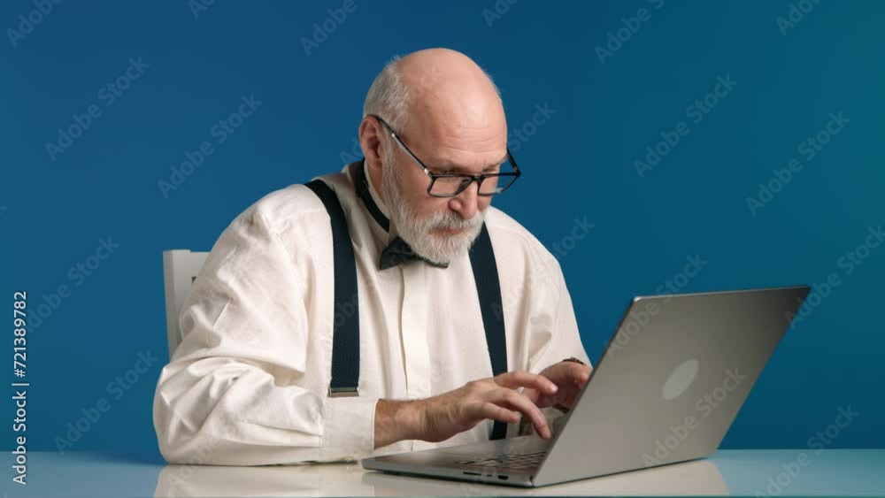 A man with a white shirt, suspenders, and bow tie is engrossed in his work on a laptop, set against a vibrant blue backdrop that highlights his concentration. High quality 4k footage A man with a white shirt, suspenders, and bow tie is engrossed in his work on a laptop, set against a vibrant blue backdrop that highlights his concentration. High quality 4k footage