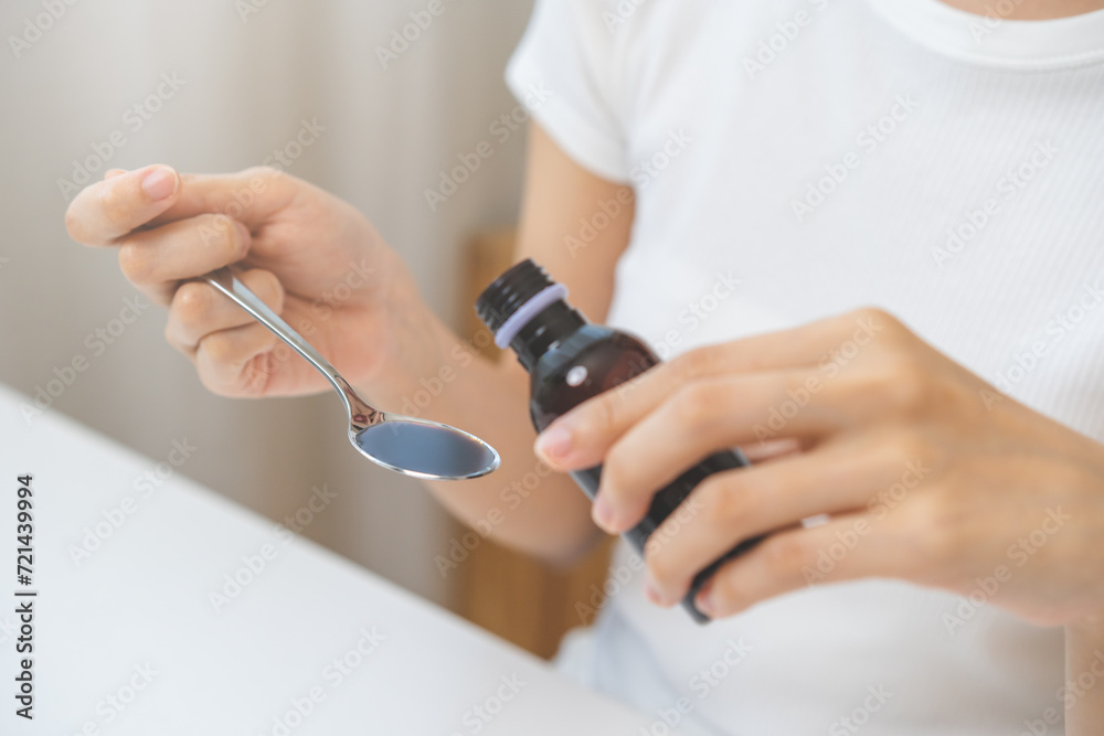 Close up hand of sick woman pouring syrup, liquid antipyretic, cough, flu medicine to stainless spoon from bottle, measuring dosage of drug with tablespoon, taking therapy, treatment cure of disease.