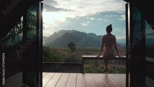 Woman relax on tropical bungalow terrace enjoy sunset mountain and jungle forest landscape. Outdoor lifestyle travel on summer vacation. Thailand, Khao Sok national park. Back view photo