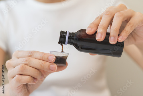 Close up hand of sick woman pouring syrup, liquid antipyretic, cough, flu medicine to jar from bottle, measuring dosage of drug with cup, taking therapy, treatment cure of disease. Health care concept photo
