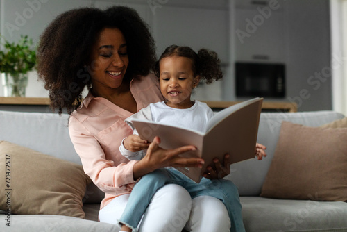 Caring young black woman reading book aloud to her cute little daughter, enjoying spending time together at home sitting on sofa photo