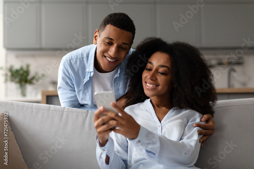 Positive African American couple sitting on couch, using smartphone, woman sharing social media content, showing photos or videos to her husband