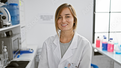 Attractive young blonde woman scientist, confidently smiling while sitting indoors, immersed in medical research in a bustling lab, portrait of confidence and joy.