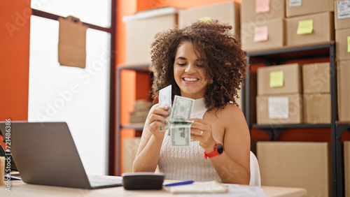 Young beautiful hispanic woman ecommerce business worker using laptop counting dollars at office photo