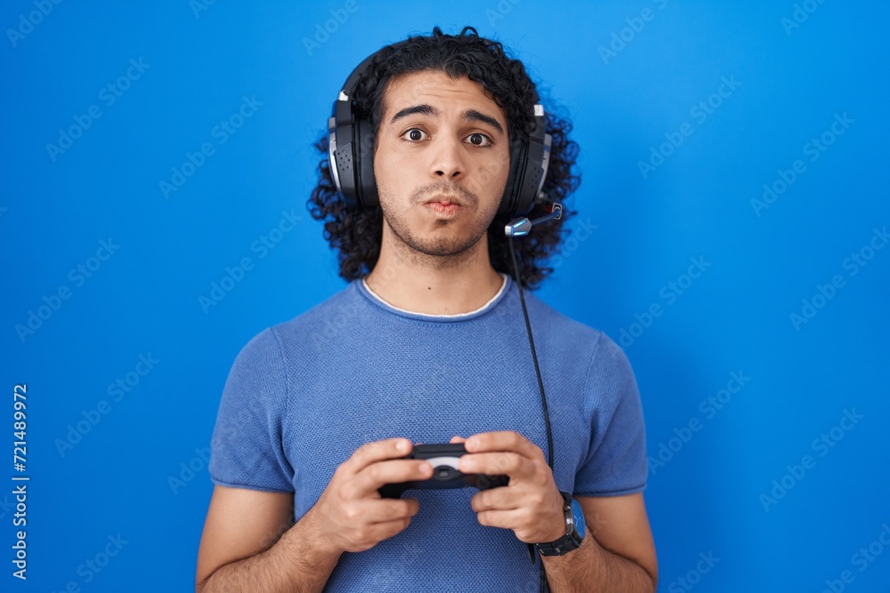 Hispanic man with curly hair playing video game holding controller puffing cheeks with funny face. mouth inflated with air, catching air. - obrazy, fototapety, plakaty 