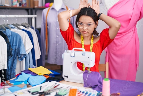 Hispanic young woman dressmaker designer using sewing machine doing bunny ears gesture with hands palms looking cynical and skeptical. easter rabbit concept. photo
