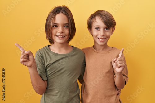 Two adorable boys standing together on a yellow isolated background, hugging and cheerfully pointing to the side with an optimistic smile.