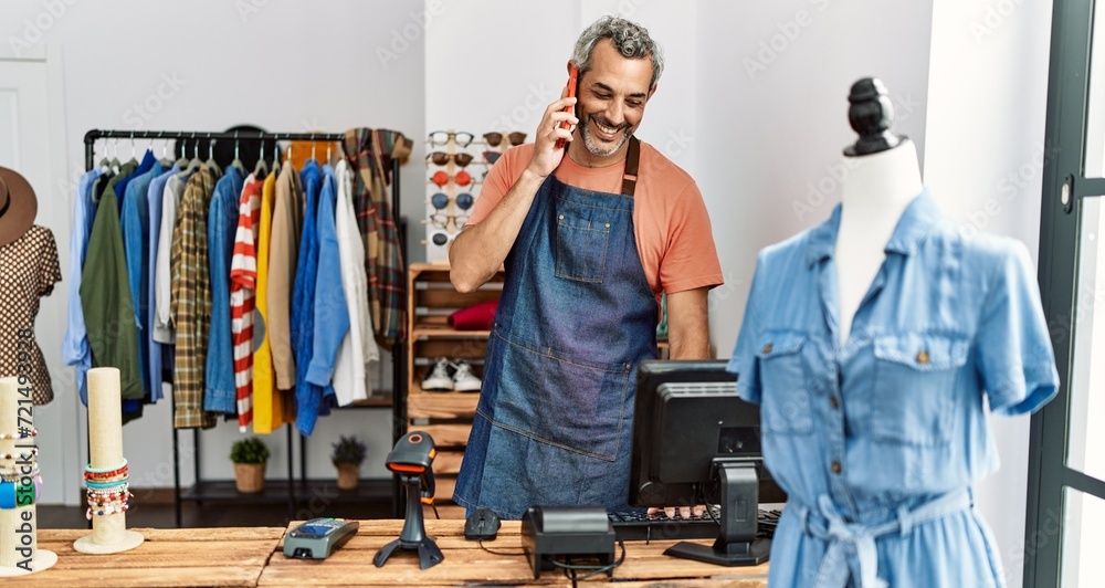 Middle age grey-haired man shop assistant using computer talking on smartphone at clothing store