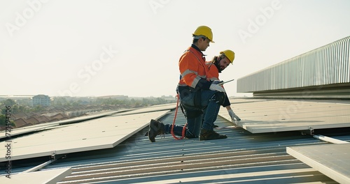 Two engineer workers in safety gear install solar panels on a rooftop, with a suburban landscape stretching out in the distance