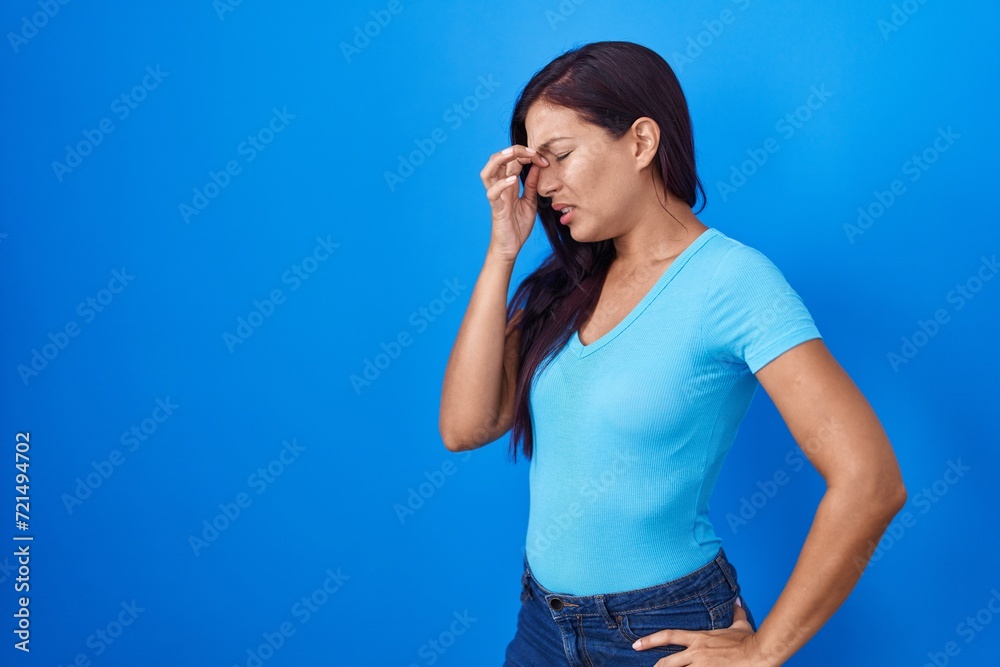 Young hispanic woman standing over blue background tired rubbing nose and eyes feeling fatigue and headache. stress and frustration concept. Young hispanic woman standing over blue background tired rubbing nose and eyes feeling fatigue and headache. stress and frustration concept.