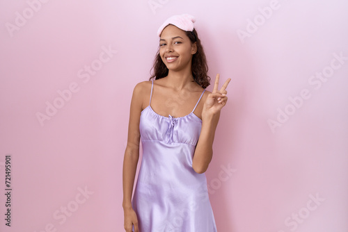 Young hispanic woman wearing sleep mask and nightgown smiling looking to the camera showing fingers doing victory sign. number two.