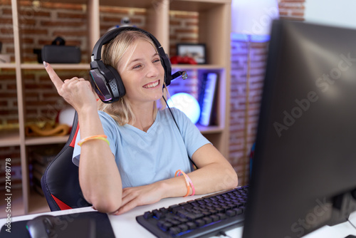 Young caucasian woman playing video games wearing headphones with a big smile on face, pointing with hand finger to the side looking at the camera.