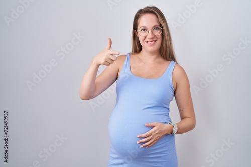 Young pregnant woman standing over white background pointing fingers to camera with happy and funny face. good energy and vibes.