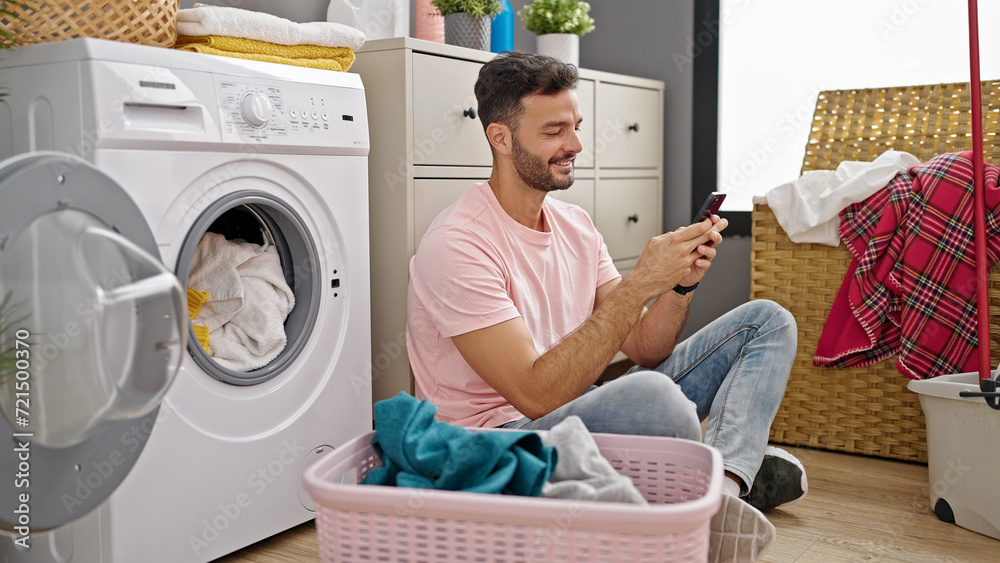 Young hispanic man using smartphone washing clothes at laundry room