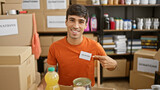 Confident hispanic young man, sitting at a table in the heart of a bustling charity center, cheerfully pointing to his volunteer badge with a big, infectious smile