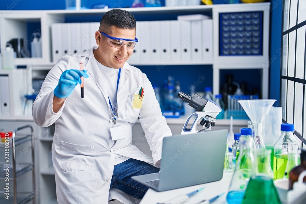 Young latin man scientist using laptop holding blood test tube at laboratory Young latin man scientist using laptop holding blood test tube at laboratory