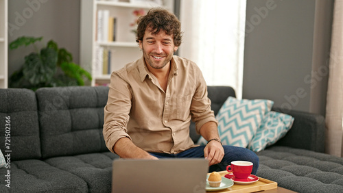 Young hispanic man having breakfast using laptop smiling at home photo