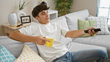 Joyful young hispanic teenager relaxing on comfortable sofa at home, sipping coffee while indulging in his favorite film on tv - just a happy guy enjoying life's little luxuries