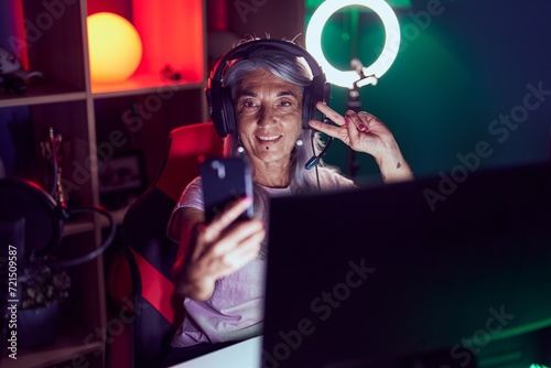 Middle age grey-haired woman streamer smiling confident make selfie by smartphone at gaming room