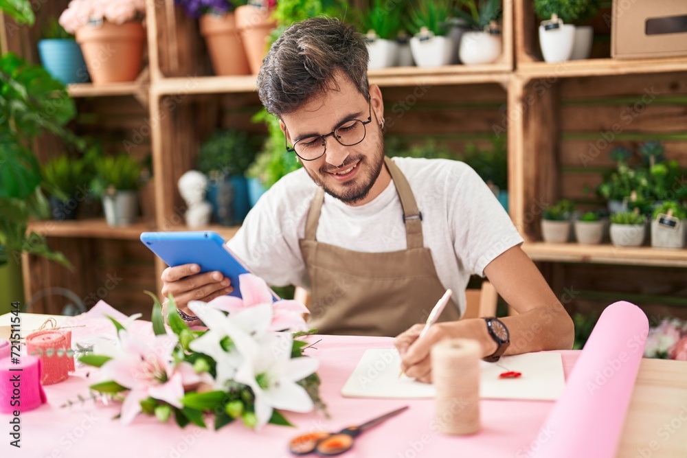 Young hispanic man florist using touchpad writing on notebook at flower shop