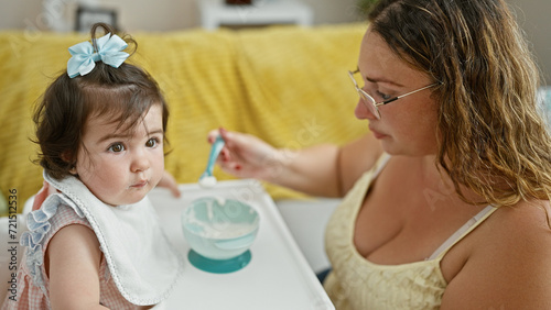 Mother and daughter together, loving family lifestyle, casually sitting at home during baby's lunch time, holding spoon and bib, concentrating on eating in the living room interior.