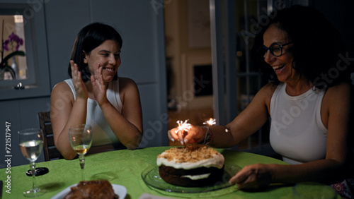 Two women  likely mother and daughter  celebrate indoors with a sparkler-topped cake and wine glasses on the table.