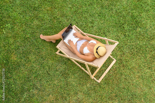 Woman in a white bikini sitting on deck chair with laptop on the green grass sunbathes at summer day. Top view, drone, aerial view.