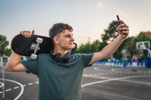 One man young adult caucasian teenager stand outdoor with skateboard on his shoulder and headphones posing at basketball court portrait happy confident wear shirt casual real person copy space photo