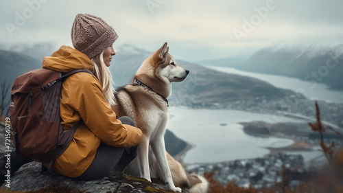 Cinematic image of a hiker girl sitting with husky at the top of the mountain with rocks, autumn trees and lake. Long shot of a beautiful scene in autumn from the top. Moody colors. © Loucine