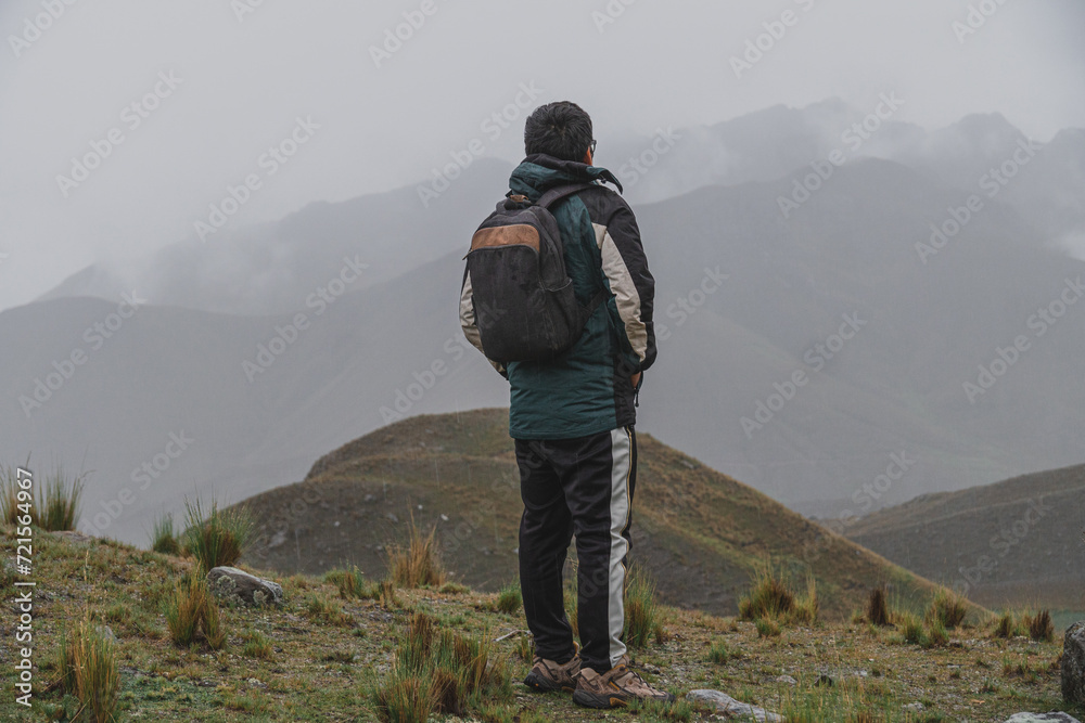 young male traveler dressed in green rain coat and black trousers, on top of mountain with panoramic view, in the rain looking at the horizon young male traveler dressed in green rain coat and black trousers, on top of mountain with panoramic view, in the rain looking at the horizon