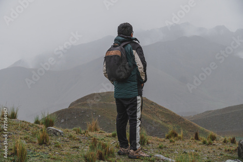 young male traveler dressed in green rain coat and black trousers, on top of mountain with panoramic view, in the rain looking at the horizon  © M Grayson
