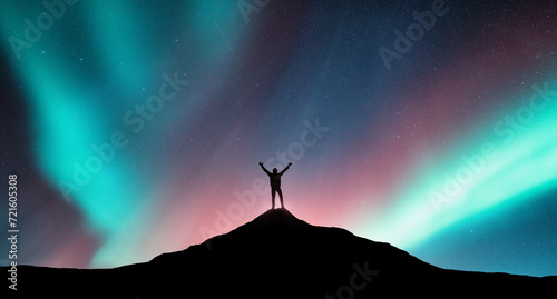 Northern lights and silhouette of a sporty man with raised up arms on the mountain peak in Norway. Aurora borealis and happy man. Sky with stars and polar lights. Night landscape with aurora. Concept