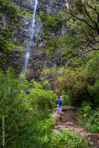 Female tourist with backpack hikes torwards a high waterfall at a rock wall. Lagoa do Vento waterfall, Madeira Island, Portugal, Europe.