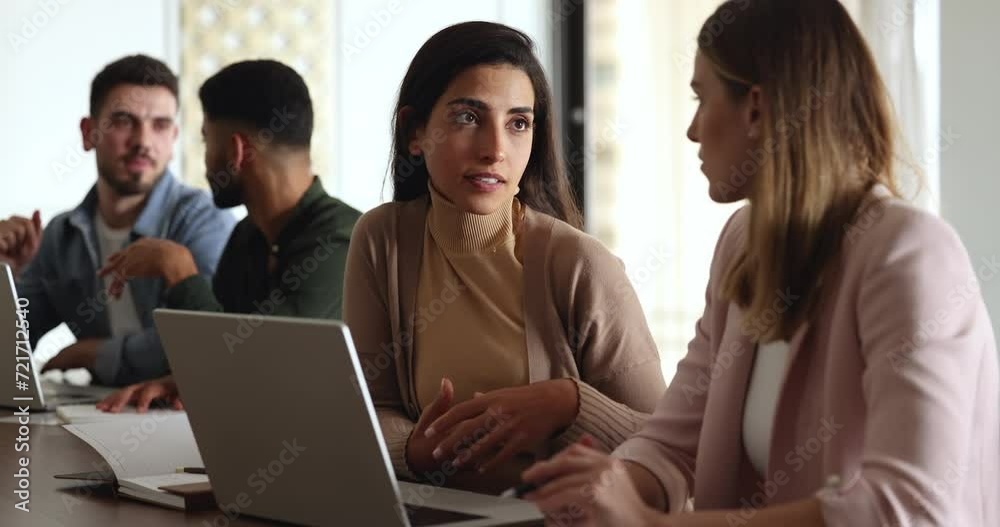 Two multiracial women colleagues sit at desk with laptop having conversation over joint collaborative, joint task. Mentor tells about corporate software to apprentice, engaged teamwork use modern tech Two multiracial women colleagues sit at desk with laptop having conversation over joint collaborative, joint task. Mentor tells about corporate software to apprentice, engaged teamwork use modern tech