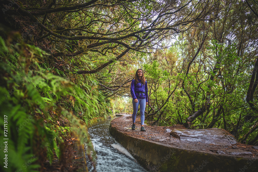 Female tourist with backpack walking along a curved, sloping water channel overgrown with laural trees on a rainy day. 25 Fontes waterfalls, Madeira Island, Portugal, Europe. Female tourist with backpack walking along a curved, sloping water channel overgrown with laural trees on a rainy day. 25 Fontes waterfalls, Madeira Island, Portugal, Europe.