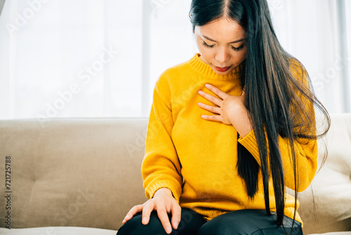 A woman from Asia suffers a heart attack on a sofa clutching her left chest in pain needing urgent medical aid. Depicting heartache discomfort and the urgency for emergency help. photo